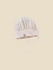 A soft, light beige baby Milton & Goose Chef's Hat with delicate stitching and fabric ties, displayed centered on a pale yellow background. The bonnet features a small tag with a logo on the side and resembles a handmade