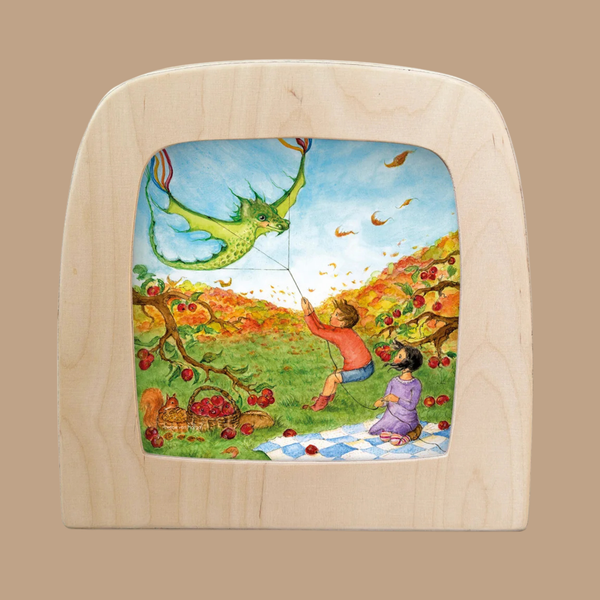 Two children sit on a picnic blanket in an autumn orchard, surrounded by apples hanging from tree branches and colorful leaves swirling in the breeze. One child flies a green dragon-shaped kite, adding a whimsical touch that reflects the enchanting spirit of the “Taming the Dragon” Silhouette for Toverlux Lamp. This silhouette-only design offers a distinctive way to bring warmth and wonder into your home decor.