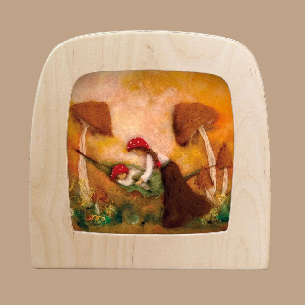 A beautifully crafted wool felt silhouette, designed for the Toverlux Lamp and framed in light wood, features red-capped mushrooms, lush green foliage, and a brown-haired figure set against an orange background. Titled “Autumn Lullaby,” this piece evokes a whimsical forest scene, with its gentle play of colors and textures capturing the enchanting charm of Magic Silhouettes.