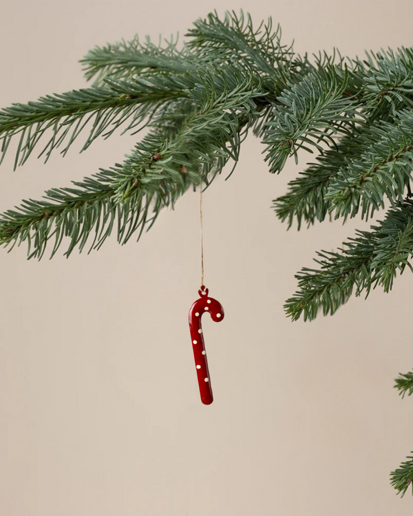 The Maileg Christmas Metal Ornament - Sugar Cane - Dot, a hand-painted red candy cane with white dots, hangs from a thread on a green pine branch against a plain beige background.