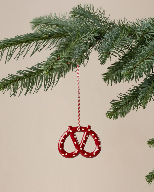 The Maileg Christmas Metal Ornament - Pretzel - Dot, a red hand-painted pretzel with white dots and a red-and-white hanging string, is displayed on a green pine branch against a plain beige background.
