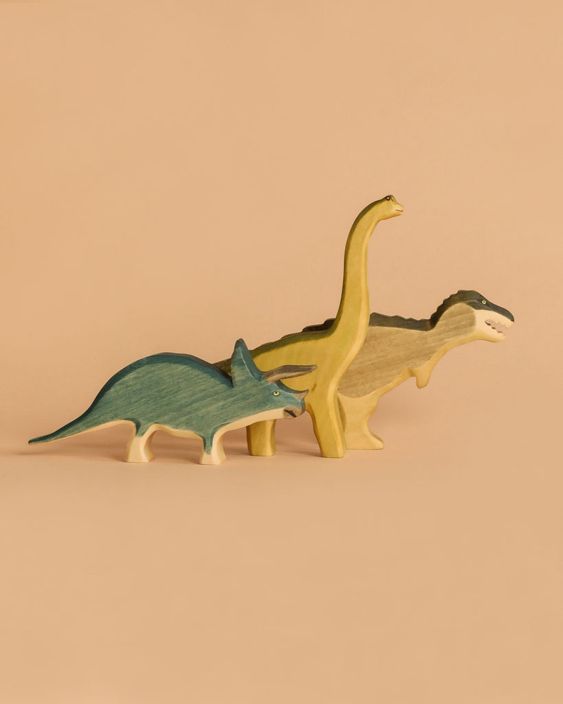 The Handmade Holzwald Dinosaur Set features a green Triceratops, yellow Brachiosaurus, and beige Tyrannosaurus rex standing together on a plain beige background, highlighting their handcrafted charm.