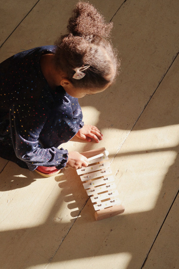 A young child with a hair clip, wearing a star-patterned shirt, plays with a Lemon wooden xylophone on a sunlit wooden floor. Shadows create a pattern around her.