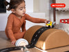Certainly! Here is the rewritten sentence incorporating the provided product name: A young child in a brown outfit engages in imaginative play, guiding a toy truck along a WaytoPlay: Small Flexible Toy Road Set - Ringroad placed on top of a brown, dome-shaped cushion. Nearby on the wooden floor are other toys and a wooden play figure.