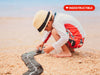A child in a hat and swimwear plays with a toy car on the WaytoPlay: Small Flexible Toy Road Set - Ringroad, which is laid out on the sandy beach. This open-ended track encourages imaginative play as a red “INDESTRUCTIBLE” label appears in the top right corner.