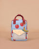 The Sticky Lemon Backpack Small - Special Edition - Apple Grub features red apple prints, red handles, a plaid front pocket with yellow flap, a green caterpillar keychain, recycled PET material, waterproof lining, and a beige background.