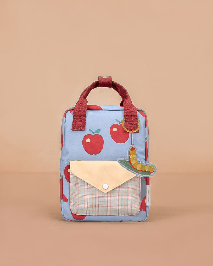 The Sticky Lemon Backpack Small - Special Edition - Apple Grub features red apple prints, red handles, a plaid front pocket with yellow flap, a green caterpillar keychain, recycled PET material, waterproof lining, and a beige background.
