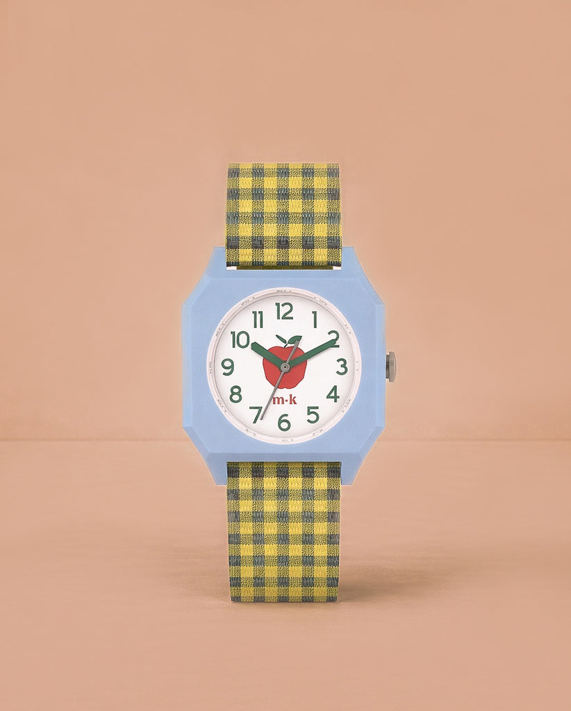 The Sticky Lemon x Mini Kyomo Watch - Apple features a light blue octagonal face, white dial with green numbers, red heart detail, and a yellow-and-black plaid strap—ideal for helping kids learn to tell time. Photographed on a plain peach background.