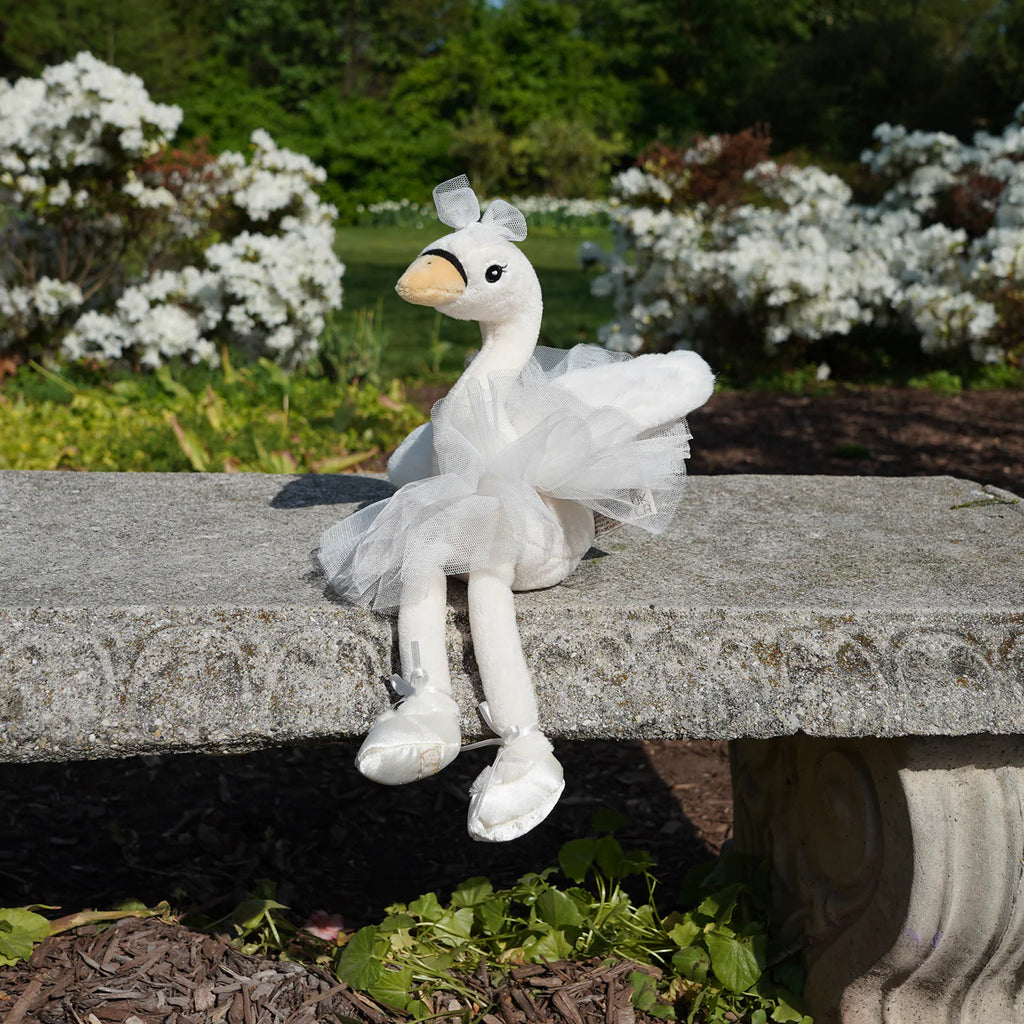 The Bukowski - Odette Ballerina Swan - White plush, dressed in a tutu and ballet slippers, sits gracefully on a stone bench amid blooming white flowers and lush greenery.