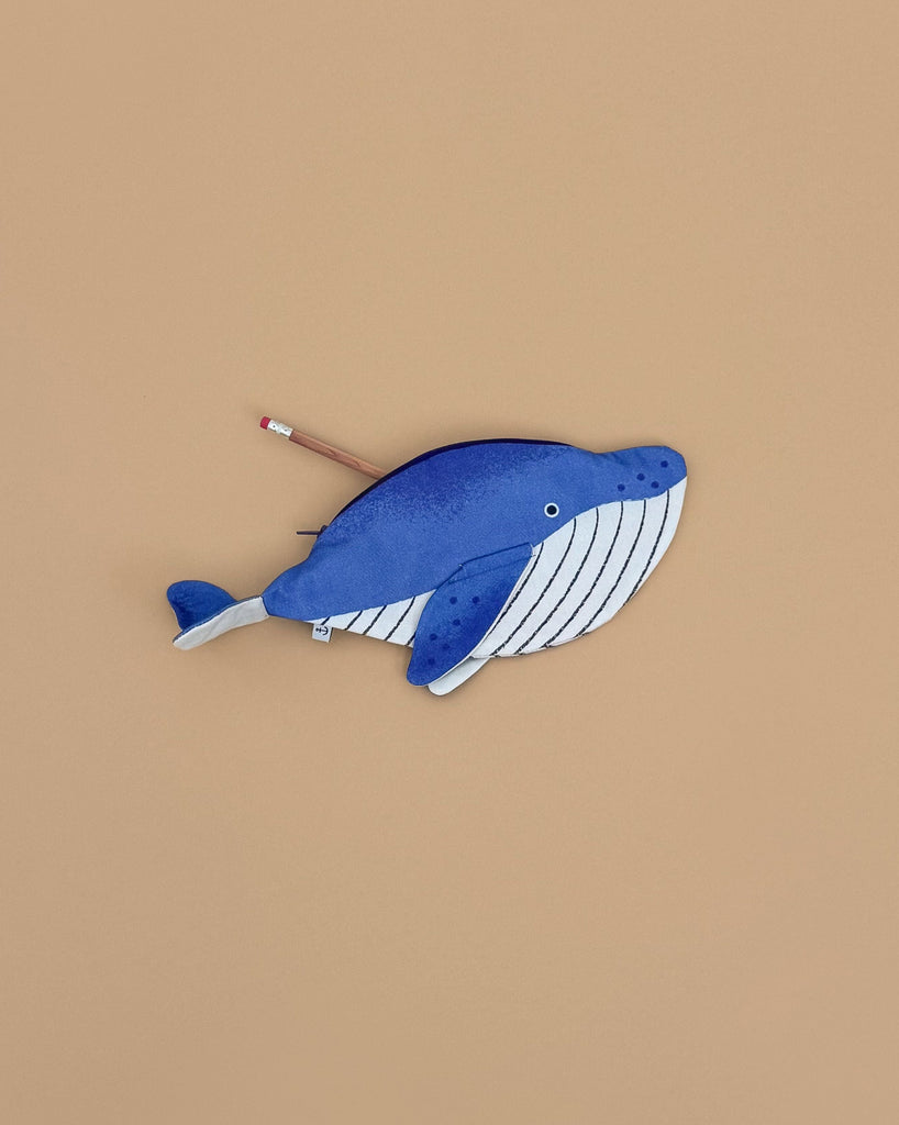 The Minke Whale Pencil Case, in blue and white and handcrafted in Spain from 100% cotton, rests on a beige surface with a pencil partly tucked into its zipper opening.