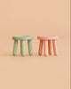 Two small Maileg stools from the Mouse series—one in light rose and the other in light mint—each crafted from beech wood with a vintage paint finish and three round legs, are placed side by side on a beige background.