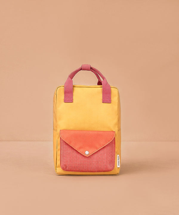 The Sticky Lemon Backpack Medium - Envelope - Apple Grub is a yellow bag with pink handles and a pink snap-button front pocket, made from recycled PET bottles, shown on a beige background.