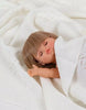 A Minikane Baby Doll With Sleeping Eyes (13") - Yzé is resting on a white knitted blanket, its brown hair complementing the serene expression of its closed eyes. One arm is gently raised, subtly showcasing its anatomically correct design, while the soft texture of the blanket partially conceals it.