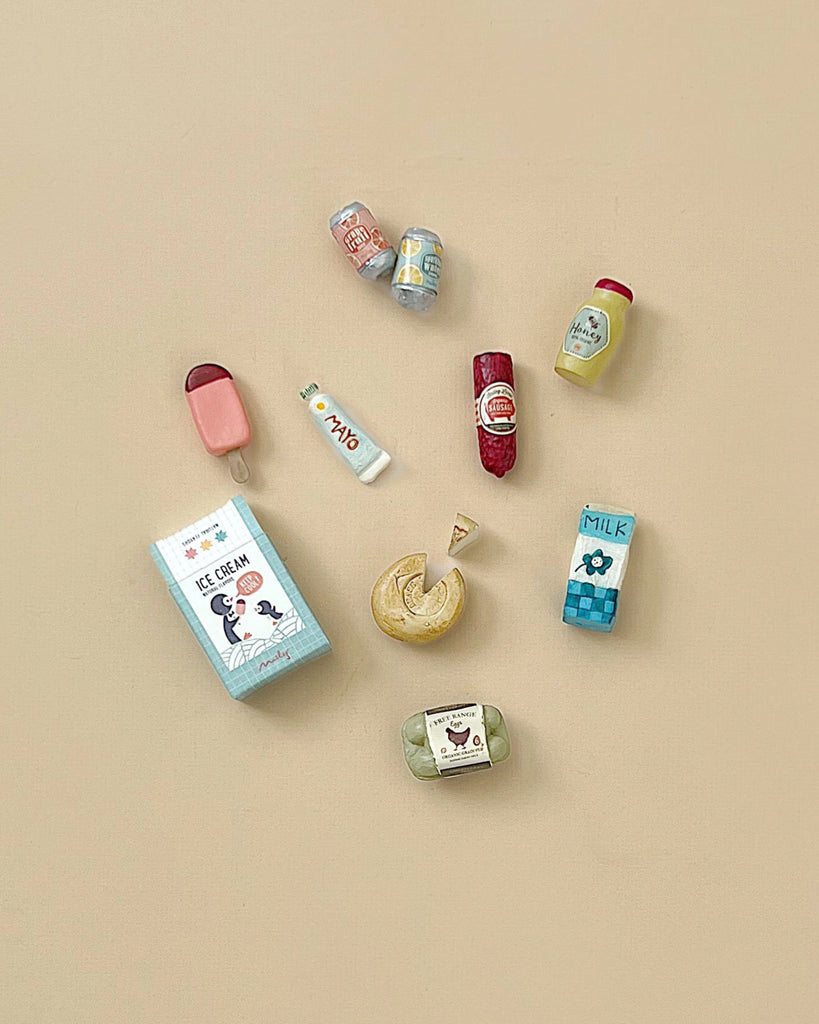 The Maileg Family Celebration Set features nine miniature food items—milk, soda cans, a popsicle, an ice cream box, a fortune cookie, a sausage, mustard, butter, and a green packaged item—artfully arranged on a beige background. Perfect as accessories for your Maileg mouse collection or for imaginative play with Maileg mice in their matchbox home.