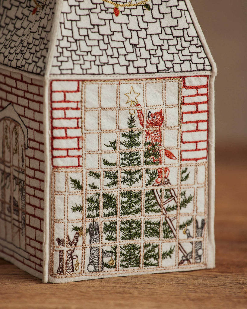 The Coral & Tusk Home for the Holidays Tissue Box Cover features an embroidered house wall with a large window, a festive tree, a red cat placing a star on top, and two smaller animals—crafted in fine detail to add holiday cheer.