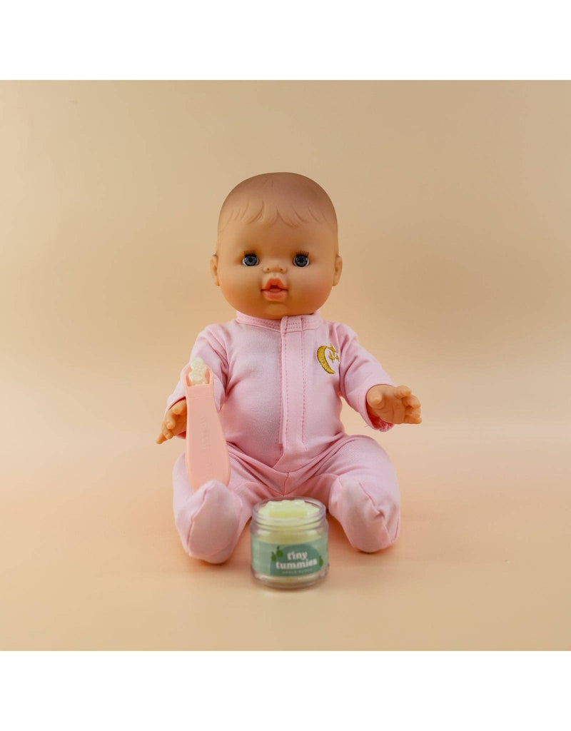 A baby doll in a pink onesie sits against a beige background, clutching a small pink bottle. In front of it is a jar labeled "Tiny Tummies - Apple jelly food" by Tiny Harlow, offering the promise of mealtime magic with every spoonful.