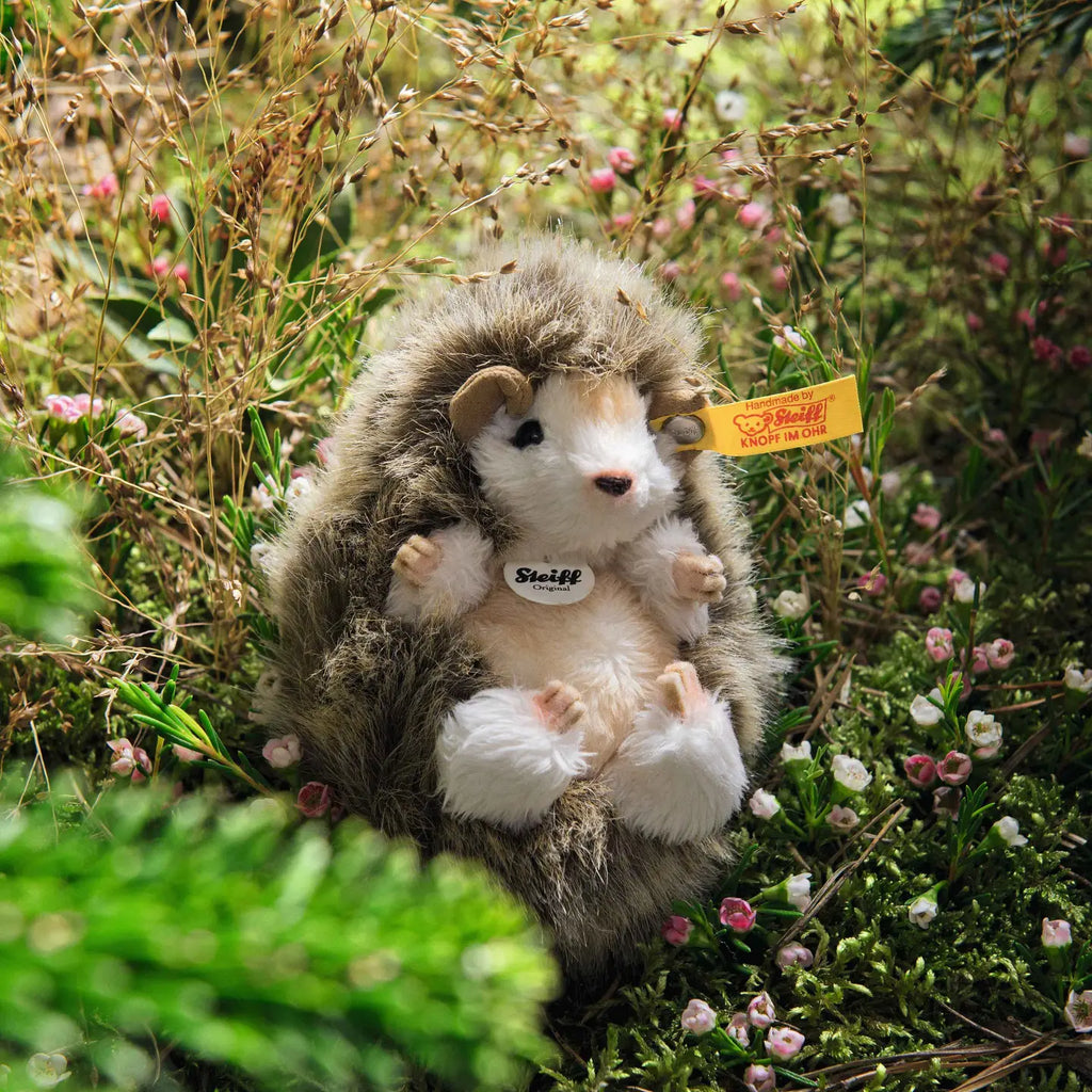 The Steiff Joggi Baby Hedgehog Stuffed Plush Toy, 4 inches, sits among green grass and small pink flowers in a natural outdoor setting.