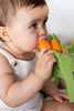 A baby with short brown hair, in a white sleeveless outfit, chews on two Cathy the Carrot Mini DouDou Teether toys while looking to the side, supporting sensory development.