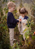 In a garden, a young boy gives a small tomato to a smiling girl holding the Senger Naturwelt Cuddly Animal - Linen Sheep plush warming pillow among lush green plants.
