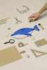 A hand reaches for a stapler among tidy beige office supplies, featuring the bright blue Minke Whale Pencil Case handcrafted in Spain at center. Glasses, scissors, ruler, notebook, tape, and clips rest on a neutral background.