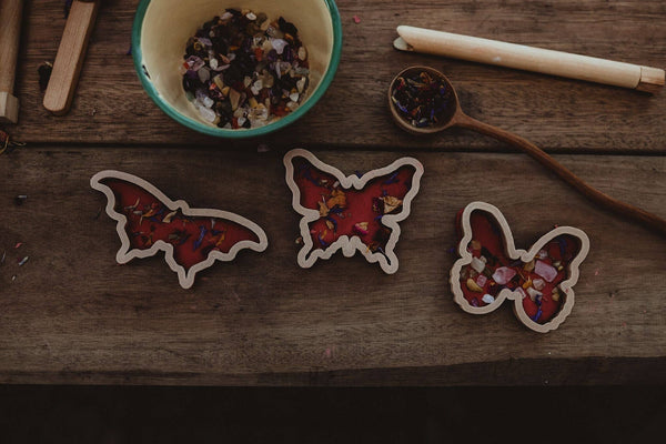 Three wooden molds from the Butterfly Cutter Set – Eco Playdough Tool are filled with red wax and dried flowers. They rest on a rustic table next to a bowl of dried flowers, a spoon, and two candle sticks—inviting sensory play with these beautiful, eco-friendly playdough cutters.