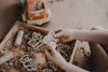 A child’s hands engage in sensory play with construction vehicle toys and sand in a wooden box, using the Construction Cutter Set - Eco Playdough Tool alongside a toy truck nearby on the floor.
