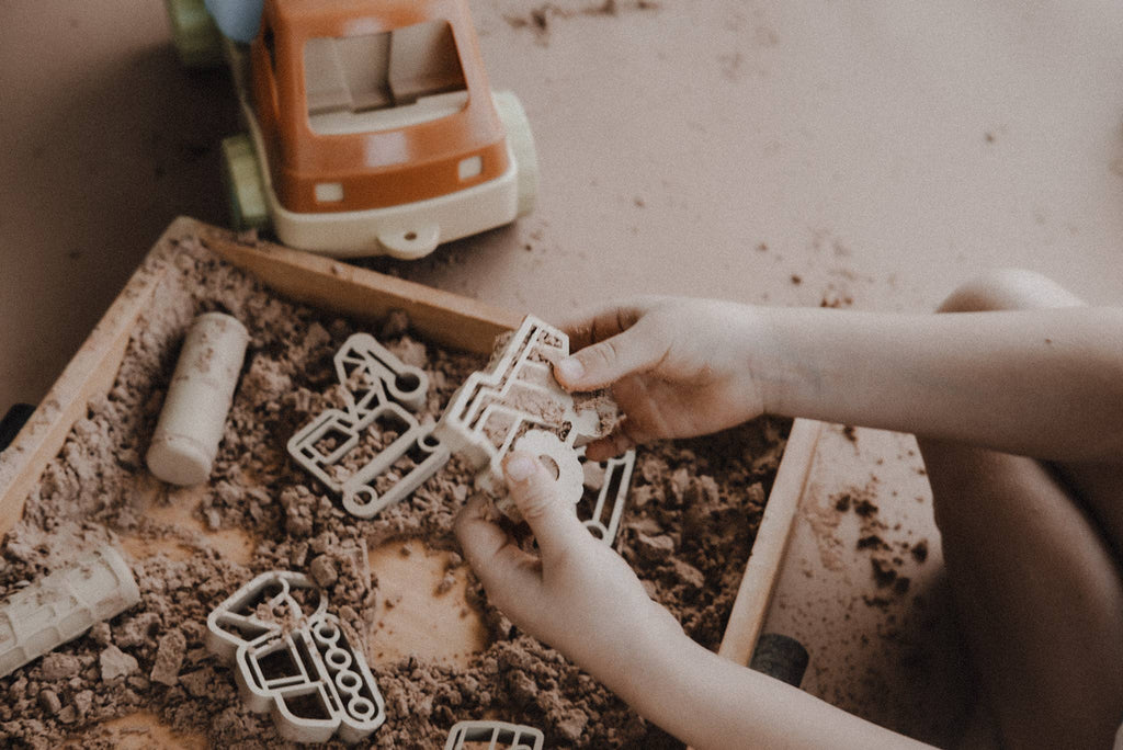 A child’s hands engage in sensory play with construction vehicle toys and sand in a wooden box, using the Construction Cutter Set - Eco Playdough Tool alongside a toy truck nearby on the floor.