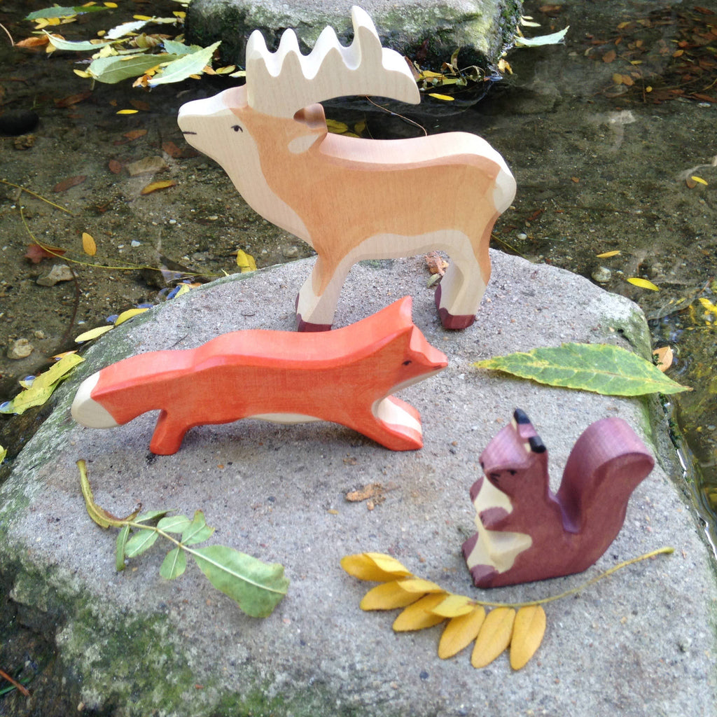 The handcrafted HOLZTIGER Squirrel, brown figure rests on a large rock near the water, surrounded by scattered green and yellow leaves.