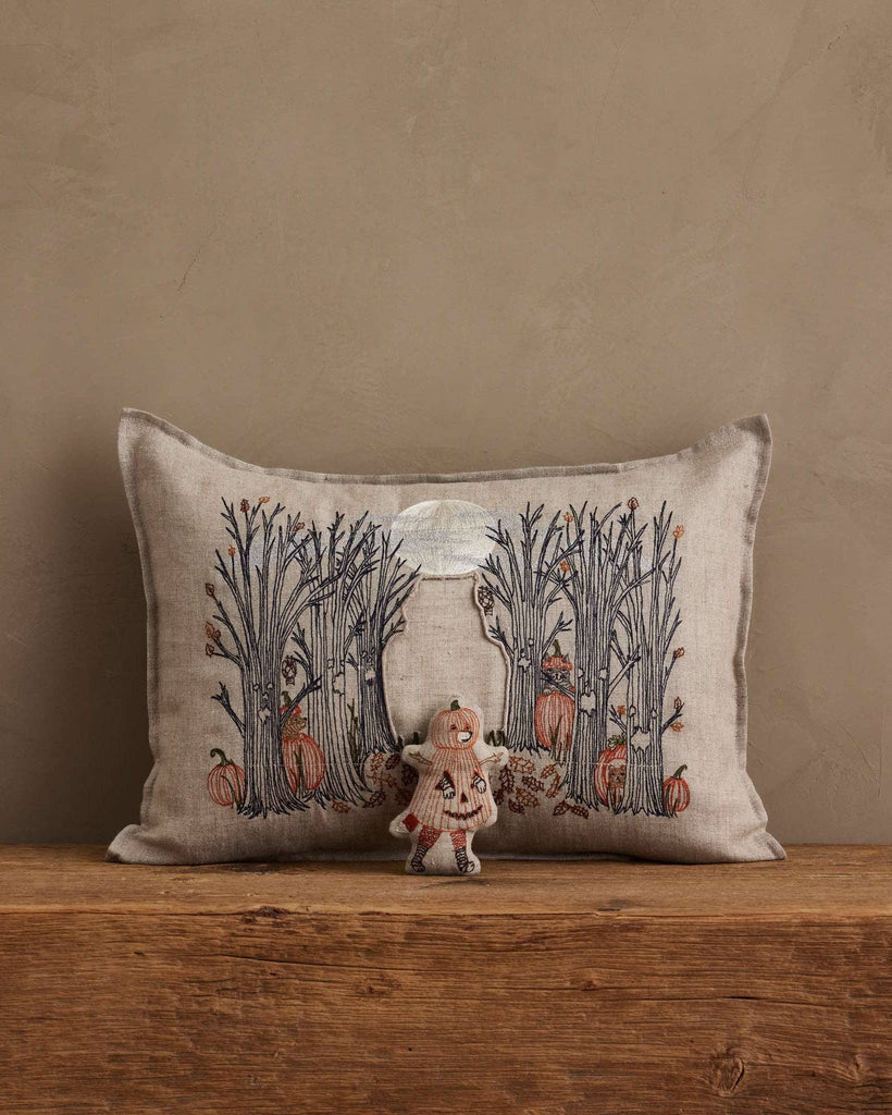 The Coral & Tusk Spooky Forest Pocket Pillow with a forest and pumpkin design rests against a beige wall, while a small pumpkin-themed doll sits in front on the wooden surface.