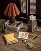 A rustic desk with a lamp, the Coral & Tusk Birdhouse Tissue Box Cover, stacked magazines, notebooks, pens, colored pencils, a frog coin purse, perfume vial, sharpener, and floral postcard in warm lighting.