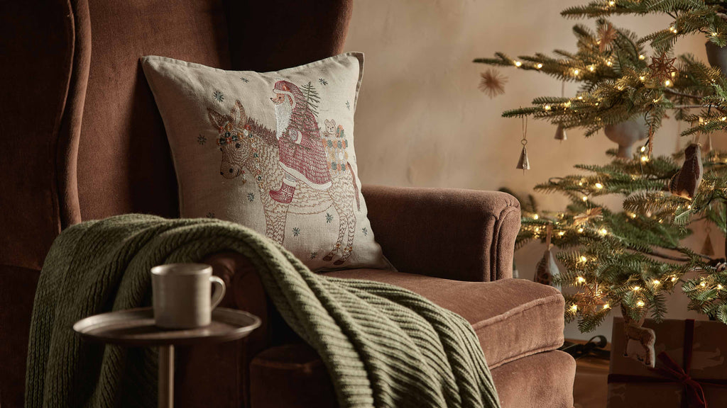 A cozy brown armchair with a green knit blanket and a Christmas Donkey Pillow sits by a small table with a mug, while a decorated Christmas tree with lights adds festive holiday charm to the warm scene.