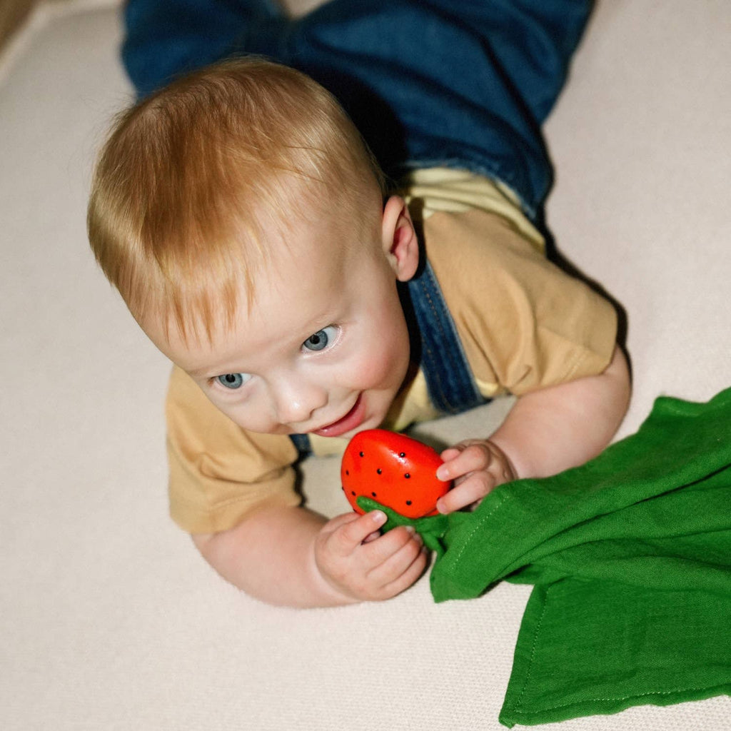 A blond baby lies on a light surface, wearing a tan shirt and denim overalls, holding Sweetie the Strawberry Big Doudou teething toy made of natural rubber and organic cotton, looking up with a curious expression.