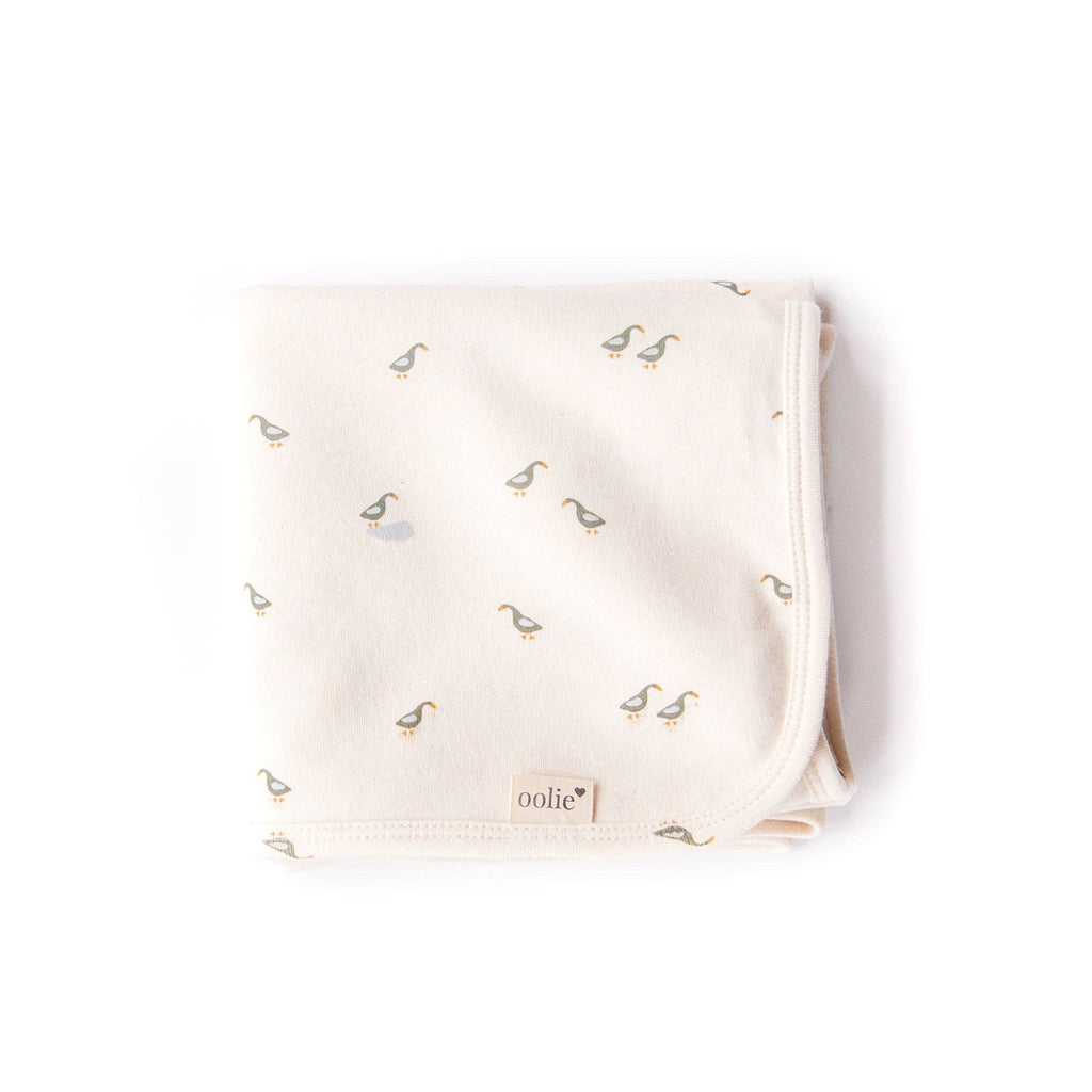 The Organic Baby Blanket – Runner Ducks is a folded, cream-colored GOTS-certified cotton blanket featuring small duck illustrations and an "oolie" fabric tag on the edge, shown on a white background for your baby's comfort and safety.