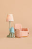 A small pink plush armchair sits beside the Maileg Stool Set, Mouse in light rose and light mint, which holds a blue teapot and a lamp with a floral lampshade, all set against a beige background.