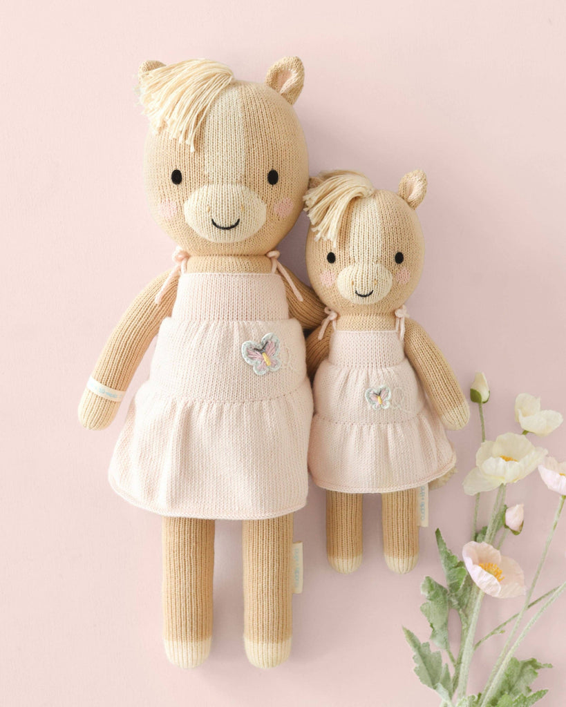 Two Cuddle + Kind Kelsie the Pony dolls, hand knit in Peru with beige yarn and blonde manes, wear matching pale pink dresses with butterfly patches. They sit on a soft pink background next to white and pink flowers.