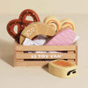 The Bakery & Patisserie Wooden Market Crate holds a charming selection of wooden pretend bread, including a pretzel, croissant, swirl pastry, pink iced pastry, and bread roll—perfect for imaginative play.