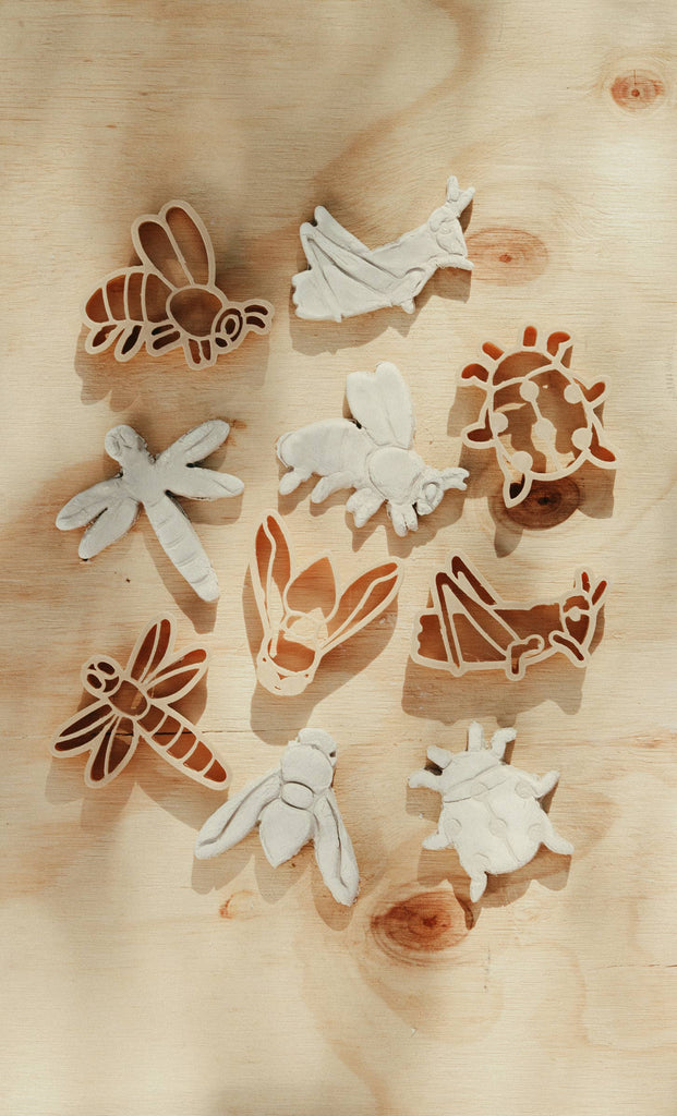Nine insect-shaped cutters from the Insect Cutter Set – Eco Playdough Tool, along with matching dough pieces, are arranged on a light wooden surface. The set features shapes like a bee, ladybug, dragonfly, moth, and grasshopper. Gentle natural light highlights these eco-friendly sensory play tools—ideal for creative playdough activities.