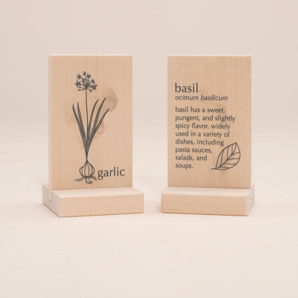 Uncle Goose Chips - Herbs features two wooden blocks made from sustainable basswood, each adorned with illustrations and text. One block highlights a garlic illustration accompanied by the word "garlic," while the other displays a basil leaf with the label "basil, ocimum basilicum" and includes information about its garden flavors and uses.