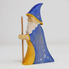 The Wooden Wizard is a hand-crafted figurine featuring a wizard with a long white beard, wearing a pointed blue hat adorned with stars and a crescent moon, and draped in a blue and yellow robe. Made from sustainable hardwood, the wizard holds a wooden staff in their right hand against a plain white background.