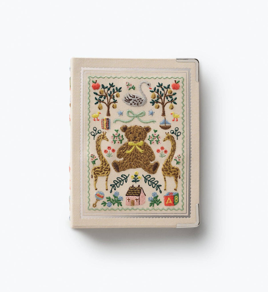The Storybook Embroidered Small Photo Album, covered in fabric and adorned with delightful embroidered motifs—including a bear, trees, swans, giraffes, flowers, a house, and alphabet blocks—graces the cover. This distinctive 4x6 album is shown against a simple white background.
