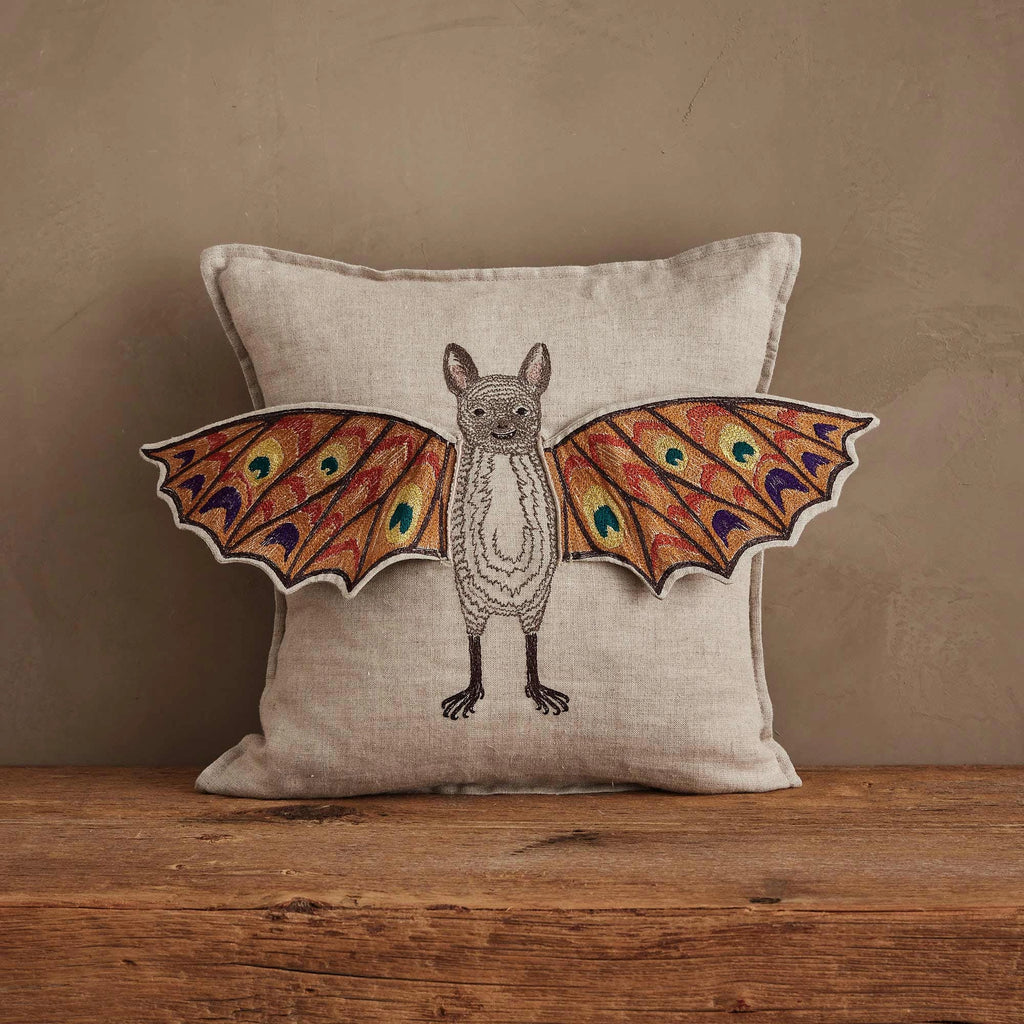 The Coral & Tusk Bat Wing Surprise Pillow showcases an embroidered bat with colorful, butterfly-like wings on a neutral backdrop, displayed on a wooden surface.