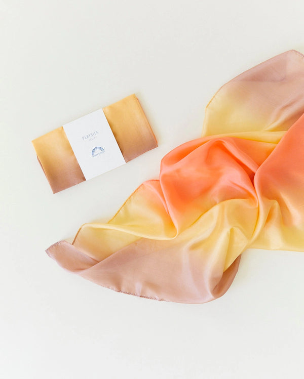 A Sarah's Silk Enchanted Playsilk in the Desert design, featuring a gradient pattern of soft brown, yellow, and orange shades, is laid out. Next to it is a folded version with a label reading "PLUSHIE." This Playsilks fabric is ideal for open-ended play with its smooth texture and luminous appearance.