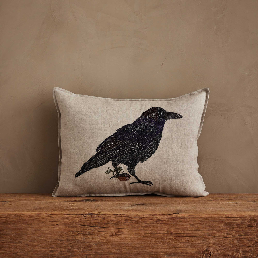 The Coral & Tusk Blackbird with Acorn Pillow is Oeko-Tex certified linen, beige in color, with an embroidered blackbird holding a branch, displayed on rustic wood against a tan wall.