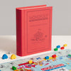 A red, fabric-bound Monopoly Junior Vintage Bookshelf Edition box stands upright next to a colorful game board with small plastic car tokens and a die.