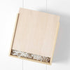 A simple light wood gift box with a sliding lid, partially open to reveal white shredded paper and the Newborn Gift Set - Pink inside, placed against a white wall background.