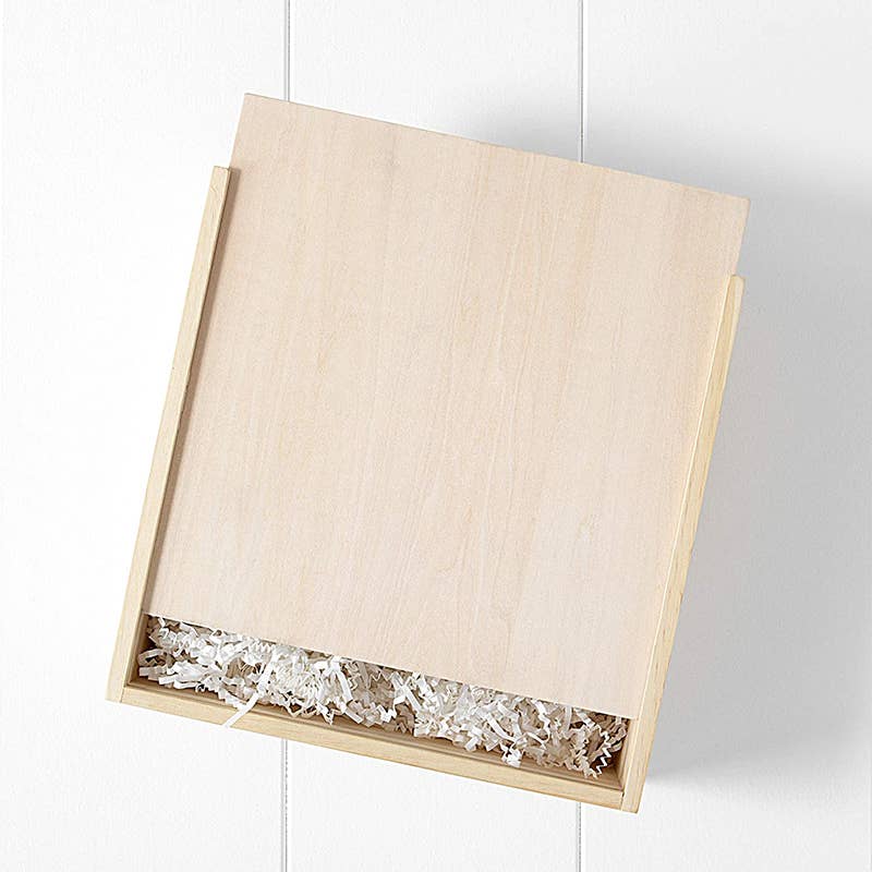 A simple light wood gift box with a sliding lid, partially open to reveal white shredded paper and the Newborn Gift Set - Pink inside, placed against a white wall background.