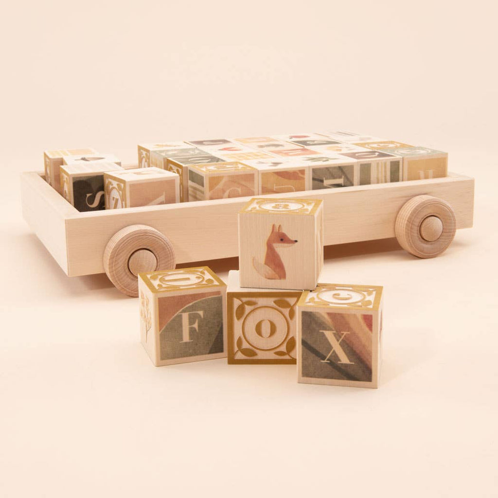 The Uncle Goose Watercolor ABC Blocks with Wagon features light-colored, patterned wooden blocks in a toy wagon. Three front blocks display F, O, X with a fox illustration—a charming and sustainable educational toy.