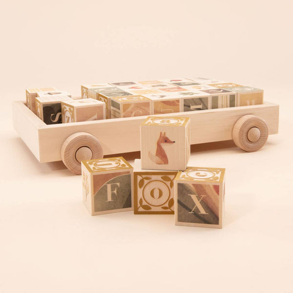The Uncle Goose Watercolor ABC Blocks with Wagon features light-colored, patterned wooden blocks in a toy wagon. Three front blocks display F, O, X with a fox illustration—a charming and sustainable educational toy.