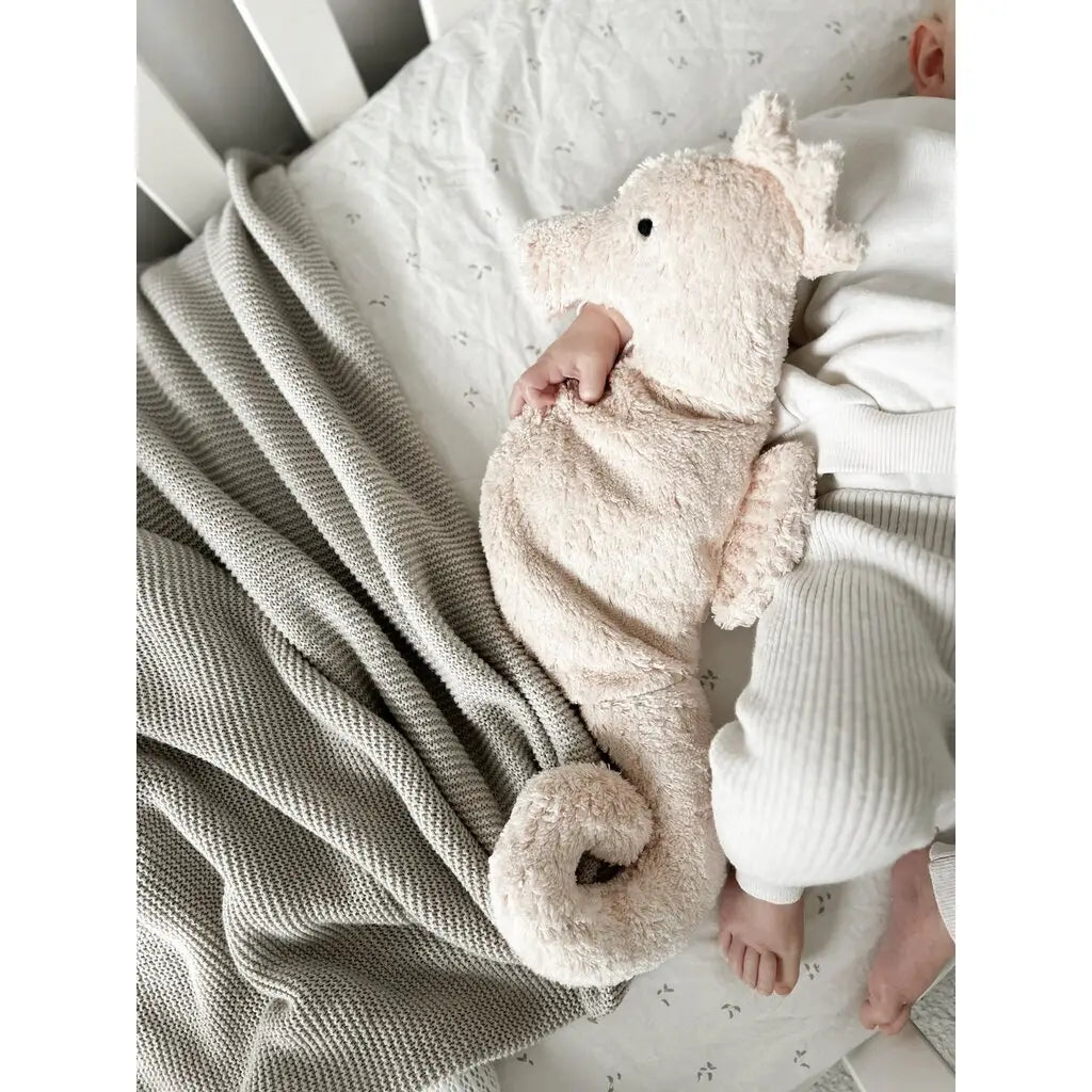 A baby cuddles the Senger Naturwelt Cuddly Animal - Seahorse while lying in a crib, dressed in white with a textured gray blanket nearby on a light-patterned bedsheet.