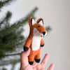 A hand holds up a Felt Finger Puppet: Fox in front of blurred green pine branches and a light background—ideal for storytelling.